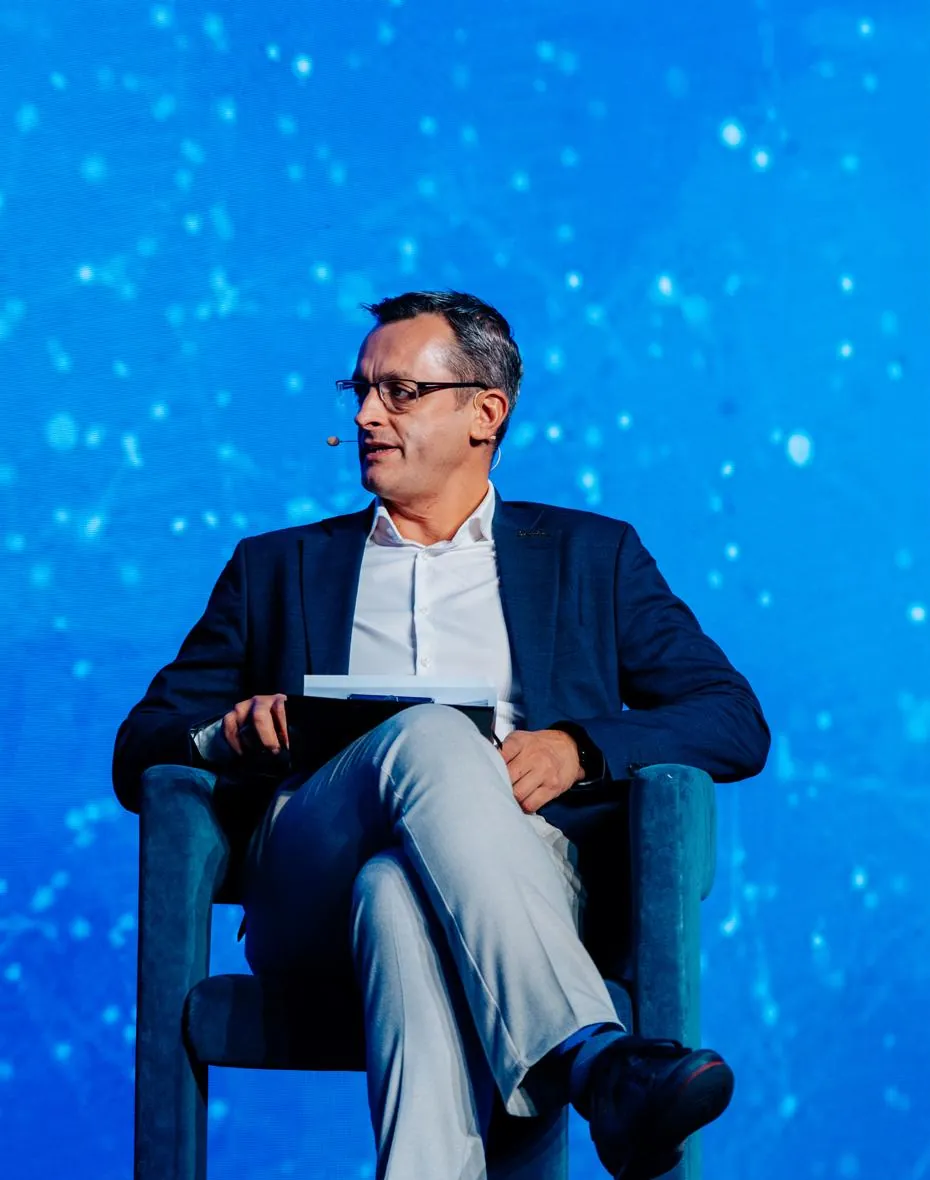 Alexander Morari, MRICS, seated on a conference stage in a navy blazer and glasses, chairing a session in front of a blue LED backdrop.