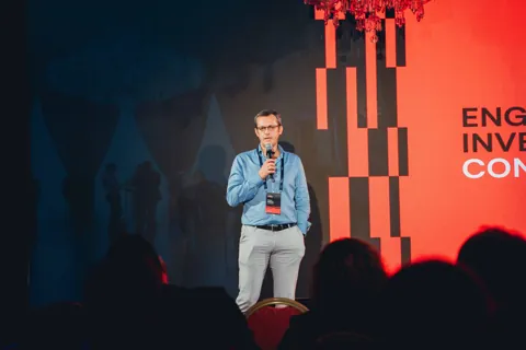 Alexander Morari alone on stage with a microphone in front of the red Engaged Investments wall in Prague, 2024.