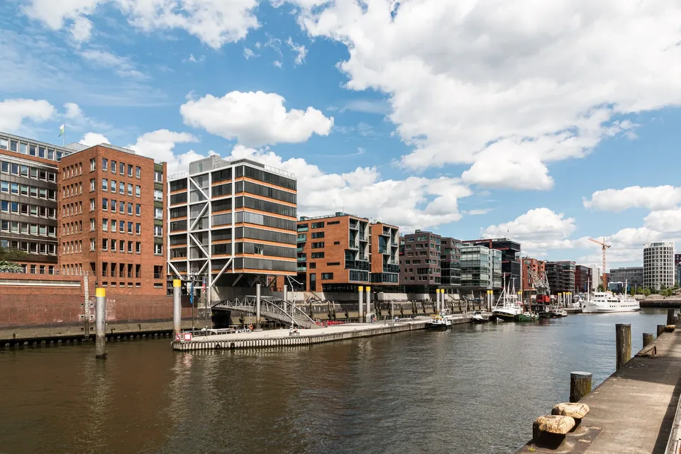 Red brick and glass residential and office blocks lining the Sandtorkai quay in Hamburg's HafenCity, reflected in the harbour basin under a bright summer sky with moored boats along the water.