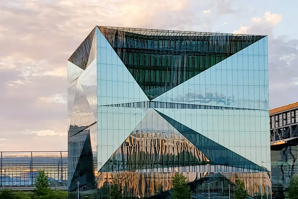 Cube Berlin, the faceted glass office building on Washingtonplatz by Berlin Hauptbahnhof, at sunset, its folded mirror façade reflecting pink evening clouds, with the station's office wing at the right edge.