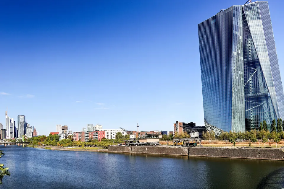 The twin glass towers of the European Central Bank in Frankfurt's Ostend rise above the River Main under a clear blue sky, with the banking district skyline in the distance.