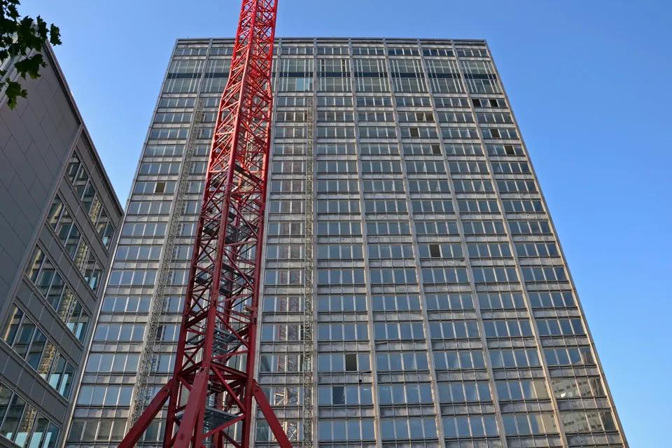 A 1960s office tower in Essen during its conversion into the Fakt Tower, seen from below in late-afternoon sun: the weathered glass curtain wall is being stripped, with panes missing on several floors, work hoists climb the facade and a red tower crane rises in front, while a neighbouring wing on the left already wears its new facade.