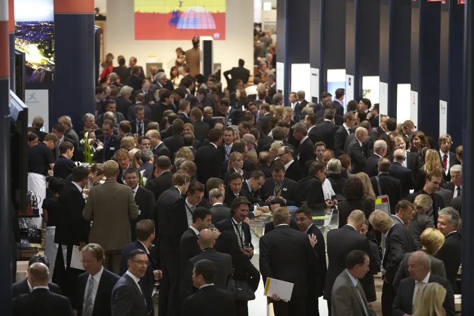 A packed hall floor at EXPO REAL 2009: hundreds of delegates in dark suits crowd around the Hamburg joint stand, talking in small groups over a long standing table.