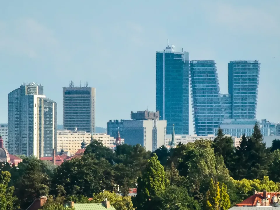 The Pankrác high-rise cluster in Prague 4 on a clear summer evening, seen over a broad band of treetops: an older glass tower & the Panorama Hotel on the left, the glass City Tower & the twisted twin blocks of V Tower on the right.