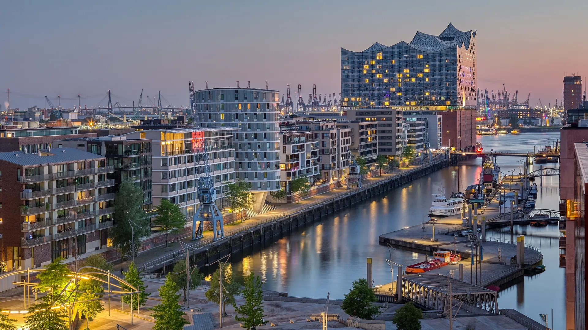Hamburg's HafenCity at blue hour: new office and residential blocks along the Sandtorhafen basin, the Elbphilharmonie behind them and the port's container cranes on the horizon.