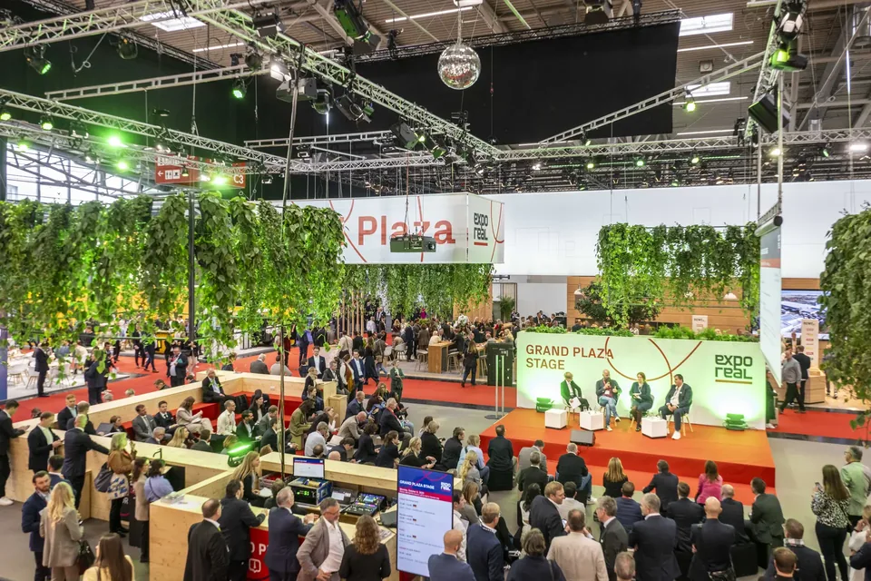 Wide view over the Grand Plaza Stage at EXPO REAL 2025: a panel under green light and hanging plants, a large audience around it and visitors crossing the hall.