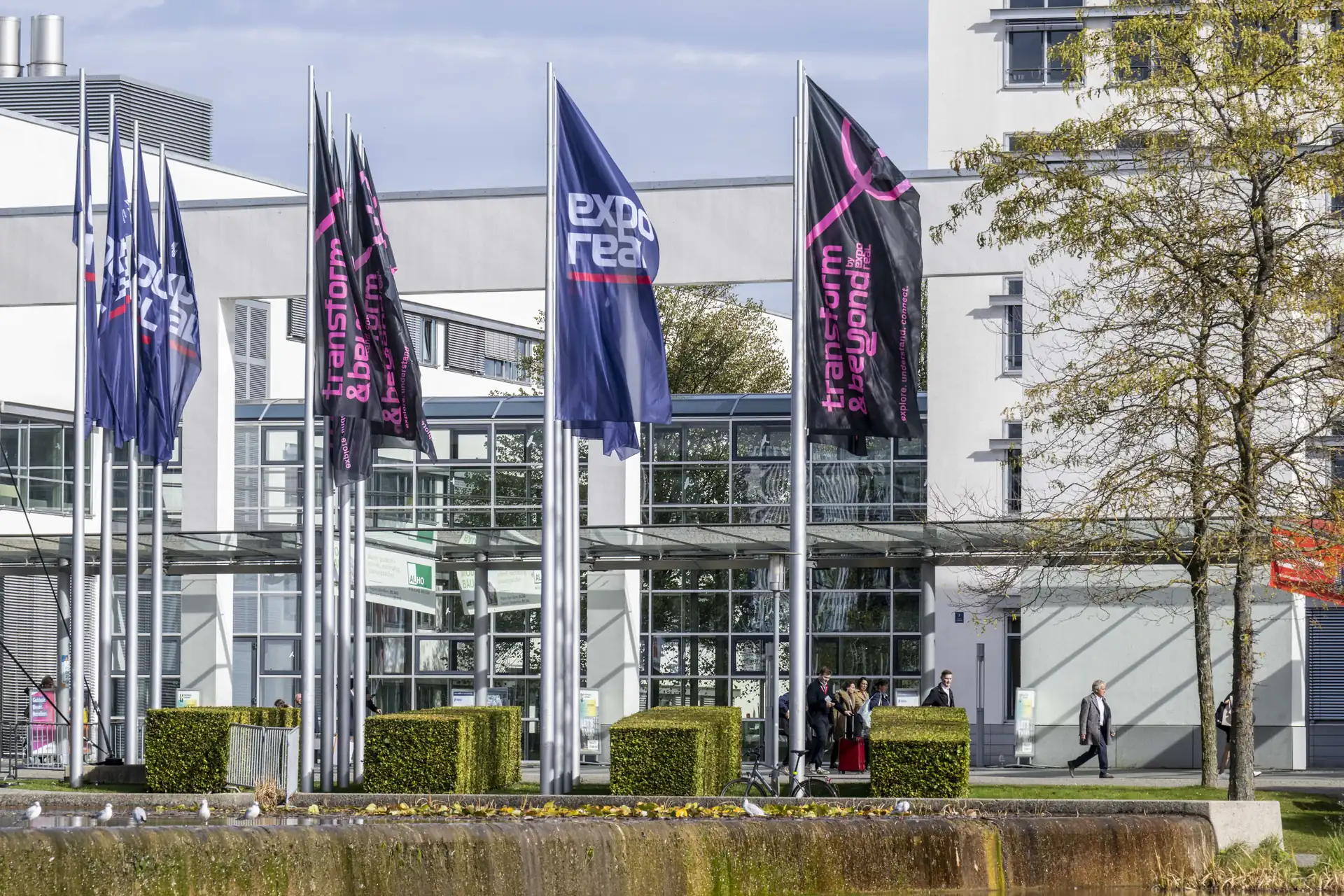 Flags outside the west entrance of Messe München during EXPO REAL 2025, with the glass entrance hall and a few visitors walking past.