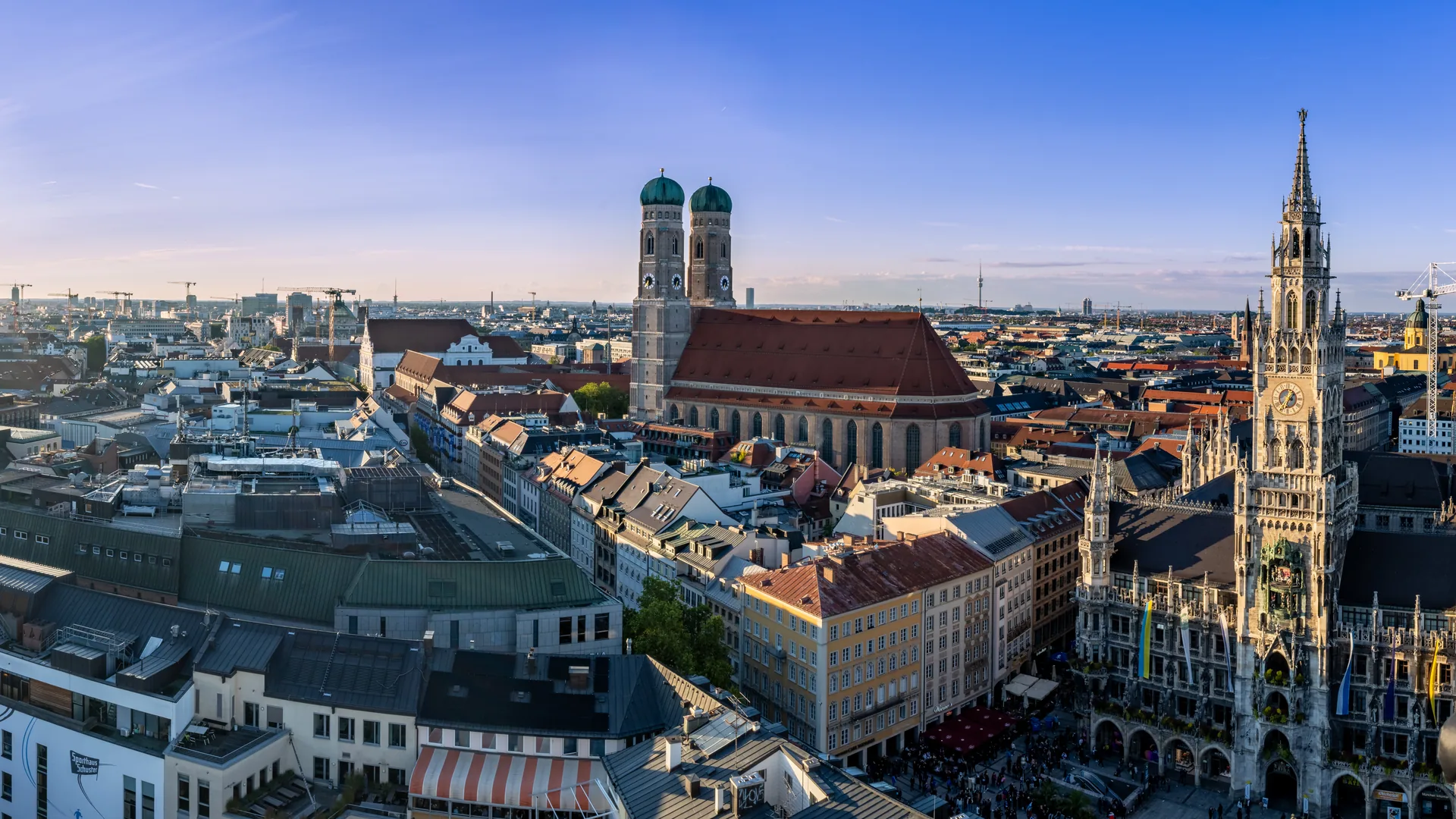 Early-evening view over central Munich from the Alter Peter tower: the twin domed towers of the Frauenkirche and the Gothic spire of the New Town Hall above the old-town roofs.