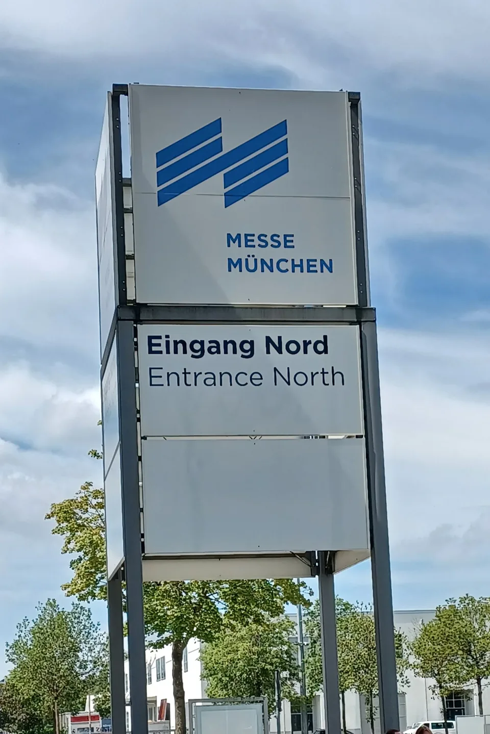 Signpost at the north entrance of the Messe München exhibition grounds, reading Eingang Nord, Entrance North, against a cloudy sky.