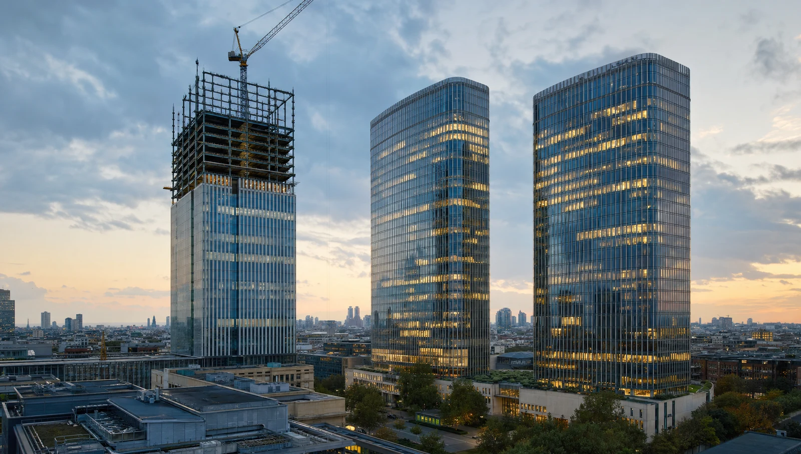 A glass office tower topping out under a crane beside two completed towers at dusk