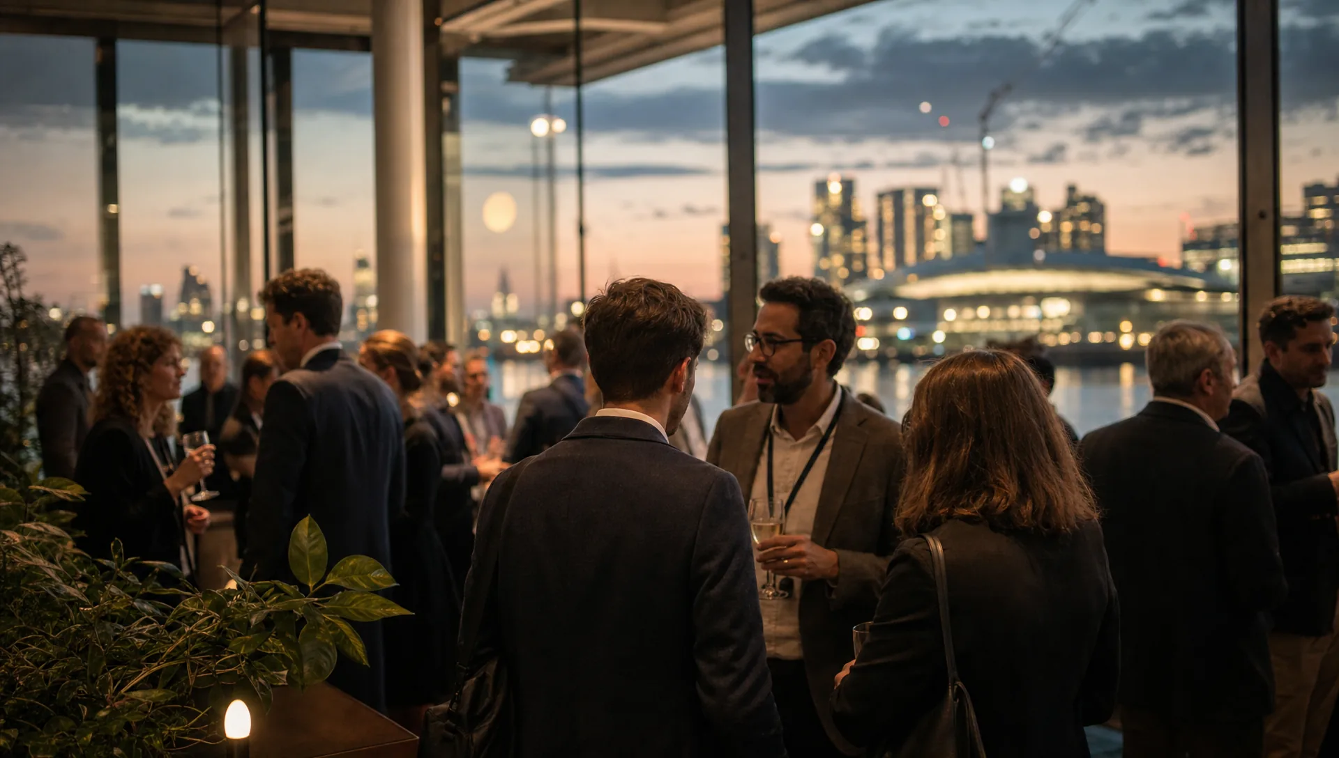 A networking reception in a glass-walled riverside venue on the Greenwich Peninsula, guests seen in profile at shallow focus.